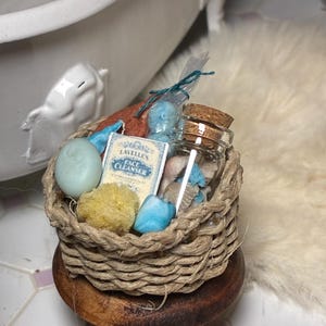 May include: A miniature woven basket filled with bath and beauty products. The basket contains a small bottle with a cork stopper, a bar of soap, a yellow sponge, and a small box of "LAVELLE'S FACE CLEANSER". The basket sits on a small wooden stool.