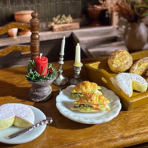 May include: Miniature food items on a wooden table. A red candle sits in a decorative holder, next to two silver candlesticks. A tray holds bread, croissants, and cheese. A plate with a slice of cheese and a knife is in the foreground.