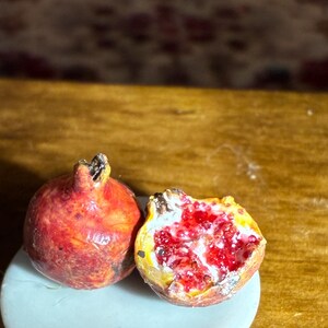 May include: Two miniature pomegranates, one whole and one halved, rest on a small, light blue plate. The whole pomegranate is a deep red with a textured surface. The halved fruit reveals bright red seeds and a yellow interior.