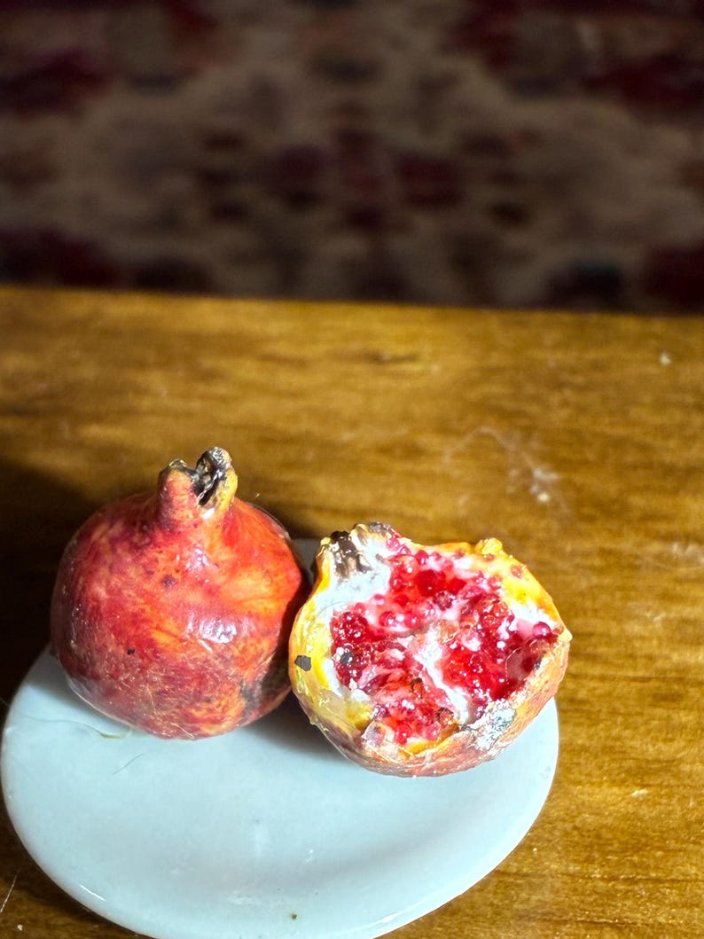 May include: Two miniature pomegranates, one whole and one halved, rest on a small, light blue plate. The whole pomegranate is a deep red with a textured surface. The halved fruit reveals bright red seeds and a yellow interior.