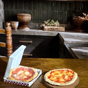 May include: Miniature pizzas, one in a checkered box and the other on a wooden serving board, sit on a wooden table. The pizzas have a red sauce base with visible toppings. The background features a kitchen setting with shelves, bowls, and dark green tile.