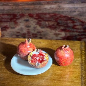 May include: Miniature pomegranates, one halved to reveal the red seeds, rest on a small white plate. Two whole pomegranates are also present. The scene is set on a wooden surface.