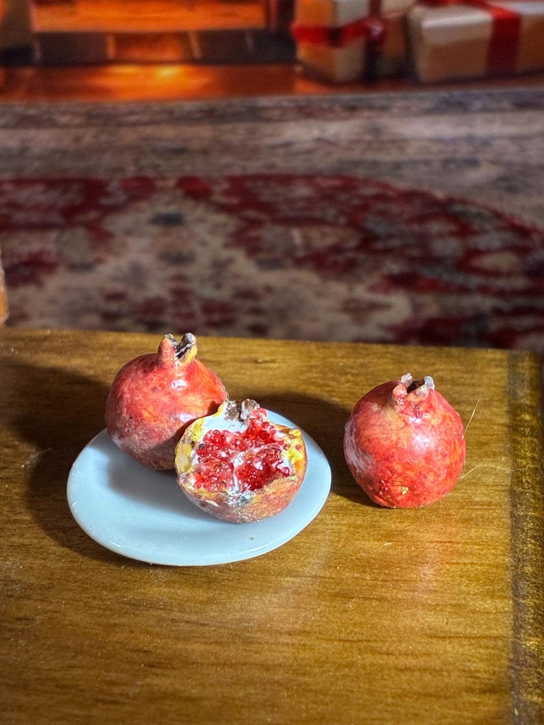 May include: Miniature pomegranates, one halved to reveal the red seeds, rest on a small white plate. Two whole pomegranates are also present. The scene is set on a wooden surface.