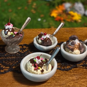May include: Miniature ice cream sundaes served in small white ceramic bowls and one clear glass pedestal dish. Each dessert features scoops of vanilla and chocolate ice cream topped with chocolate syrup, whipped cream, sprinkles, and a red cherry.