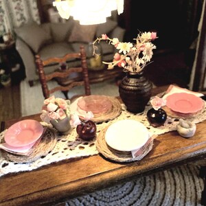 May include: A miniature dining table set for a meal. The table is made of wood and has a white lace runner. It is set with pink plates, woven placemats, and small vases with flowers. A dark vase holds a floral arrangement.