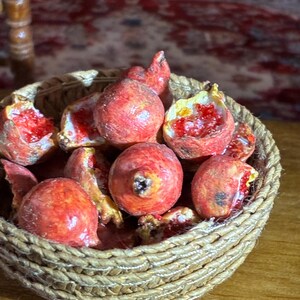 May include: A woven basket filled with miniature, realistic pomegranates. The pomegranates are a vibrant red with exposed seeds, some whole and some cut open. The basket is made of a light brown, woven material.