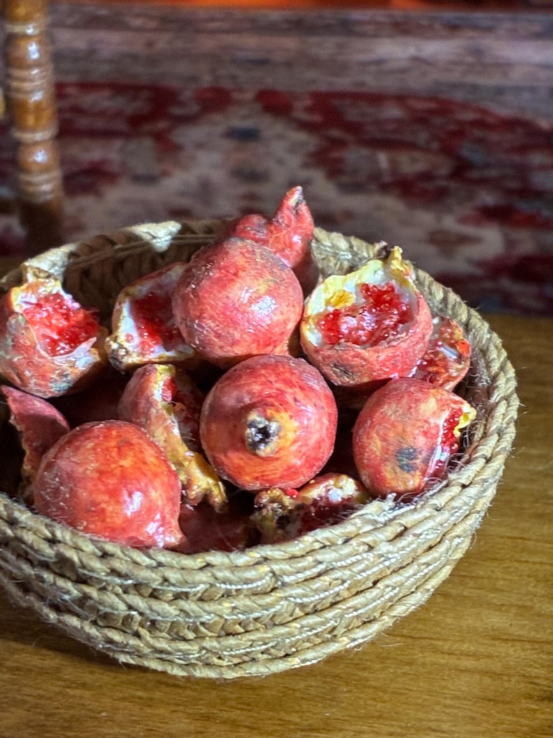 May include: A woven basket filled with miniature, realistic pomegranates. The pomegranates are a vibrant red with exposed seeds, some whole and some cut open. The basket is made of a light brown, woven material.