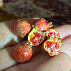 May include: Miniature, realistic pomegranates. Some are whole, others are cut open, revealing bright red seeds and white membranes. The pomegranates have a reddish-orange hue with a textured surface, and are held in a hand.