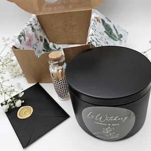 Könnte beinhalten: Ein schwarzes Kerzengefäß mit einem silbernen Etikett, auf dem "L-Witchery candles & more" steht. Das Kerzengefäß befindet sich in einer Geschenkbox mit floralem Seidenpapier. Ein kleines Glas mit Streichhölzern ist ebenfalls enthalten.