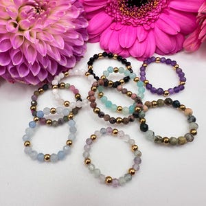 Könnte beinhalten: Eine Sammlung von Armbändern mit Perlen in verschiedenen Farben, darunter Schwarz, Blau, Rosa und Grün, mit goldfarbenen Perlen. Die Armbänder sind auf einer weißen Oberfläche angeordnet, mit rosa und lila Blumen im Hintergrund. Die Armbänder sind ein Schmuckstück.