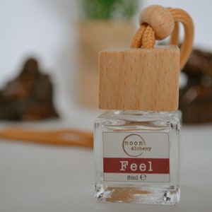 May include: A small glass bottle with a wooden cap and a brown string. The bottle has a label that says "moon alchemy" and "Feel 8ml e".