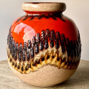 Puede incluir: Un jarrón de cerámica redondo con una superficie texturizada. La mitad superior es de color rojo brillante, que pasa a una banda texturizada negra y marrón, y termina con una base beige. El jarrón tiene una pequeña abertura en la parte superior.