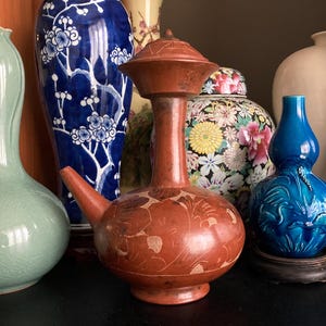 May include: A reddish-brown ceramic water vessel with a spout and lid, adorned with etched floral patterns. The vessel is part of a collection of decorative vases, including a blue and white floral vase and a turquoise gourd-shaped vase.
