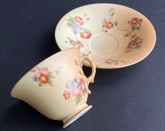 May include: A vintage teacup and saucer set. The cup and saucer are a pale cream colour with a floral design in red, pink, blue, and purple. The cup has a curved handle and sits next to the saucer on a dark surface.