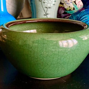 May include: A green ceramic bowl with a crackle glaze and a dark brown rim. The bowl is round and has a wide opening. Other ceramic vases are visible in the background.