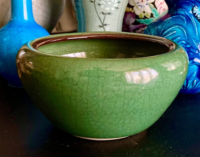 May include: A green ceramic bowl with a crackle glaze and a dark brown rim. The bowl is round and has a wide opening. Other ceramic vases are visible in the background.