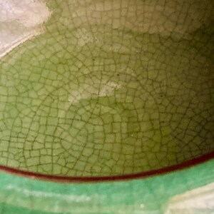 May include: Close-up of a ceramic surface with a crackle glaze in shades of green and brown. The glaze has a network of fine lines, creating a textured appearance. The rim is a darker green with a thin brown band.