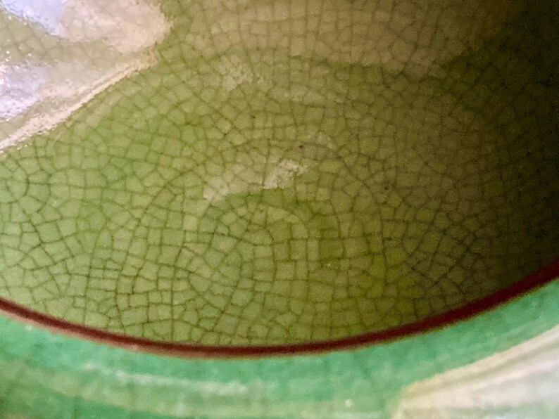 May include: Close-up of a ceramic surface with a crackle glaze in shades of green and brown. The glaze has a network of fine lines, creating a textured appearance. The rim is a darker green with a thin brown band.