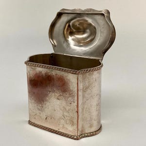 Puede incluir: Un recipiente de metal plateado con una tapa abatible unida. El recipiente rectangular presenta esquinas redondeadas, un borde estilo cuerda en los bordes superior e inferior y una superficie interior pulida. El exterior muestra signos de deslustre.