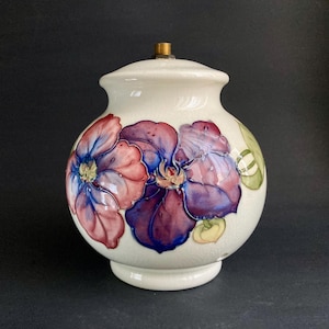 May include: A cream-coloured ceramic lamp base with a lid, decorated with hand-painted flowers in purple, red, and blue. The lamp has a brass fitting and a round base. The floral design includes green leaves and yellow accents.