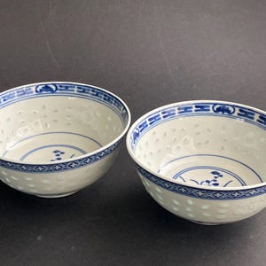 May include: Two white ceramic bowls featuring a translucent rice grain pattern throughout the body. Each bowl is decorated with a blue geometric border along the rim and a small blue floral motif centred at the bottom of the interior basin.