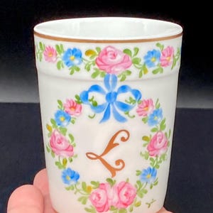 May include: A white porcelain tumbler featuring a hand-painted floral wreath of pink roses and blue forget-me-nots. A blue ribbon is tied at the top of the wreath, which encircles a gold-colored letter L. The rim and base have thin gold trim.