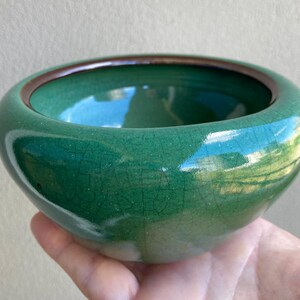 May include: A small, round, green ceramic bowl with a dark brown rim. The bowl has a glossy finish with a crackle effect. The bowl is held in a hand.