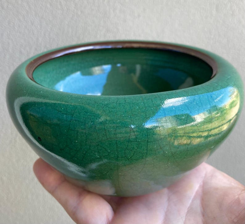 May include: A small, round, green ceramic bowl with a dark brown rim. The bowl has a glossy finish with a crackle effect. The bowl is held in a hand.