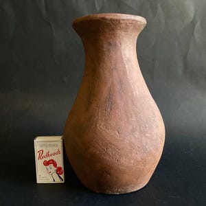 Può includere: Un vaso alto in terracotta marrone, con una base ampia e un bordo svasato. Il vaso ha un aspetto rustico e fatto a mano. Una scatola di fiammiferi "Redheads" è accanto al vaso.