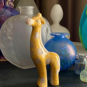 Puede incluir: Una figura de jirafa de cerámica amarilla con marcas cuadradas marrones. La jirafa tiene un cuello largo y está de pie. Varias jarrones de vidrio decorativos de varias formas y colores están en el fondo.