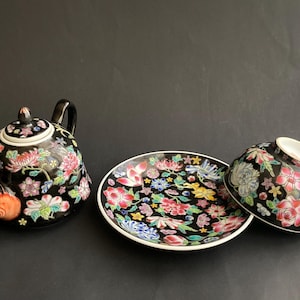 Puede incluir: Un juego de té de cerámica negra que incluye una tetera, un platillo y una taza. Cada pieza está decorada con un vibrante patrón floral multicolor que incluye flores rosas, rojas, azules y amarillas sobre un fondo negro brillante.