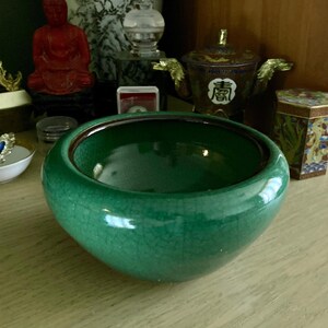 May include: A round, emerald green ceramic bowl with a crackle glaze. The bowl has a dark brown rim and sits on a light-colored wooden surface. Other decorative items are visible in the background, including a small dish with jewelry.