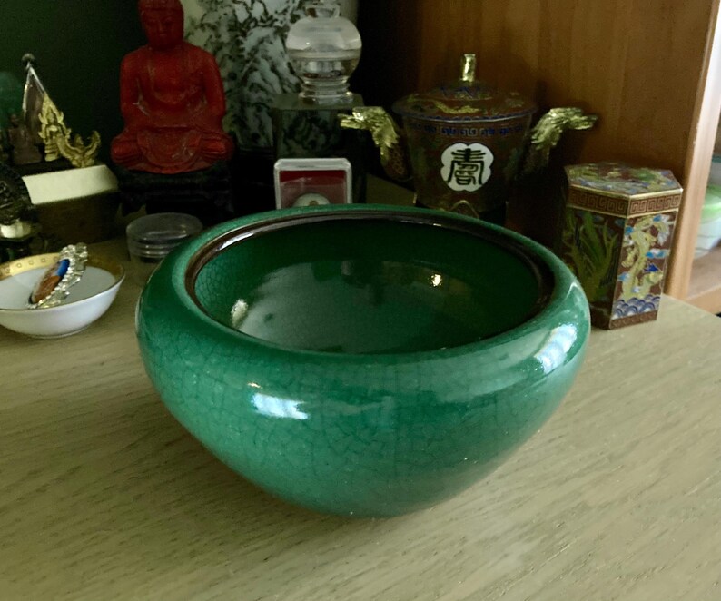May include: A round, emerald green ceramic bowl with a crackle glaze. The bowl has a dark brown rim and sits on a light-colored wooden surface. Other decorative items are visible in the background, including a small dish with jewelry.
