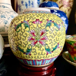 May include: A yellow ceramic jar with a floral design. The jar has a rounded shape and is decorated with pink, green, and blue floral patterns. It sits on a red wooden stand, with other decorative vases in the background.