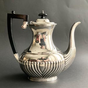 Peut inclure: Une théière argentée polie avec une poignée et un bouton de couvercle noirs. La théière a un bec incurvé et un motif décoratif nervuré sur la partie inférieure. La surface réfléchissante reflète l'environnement.