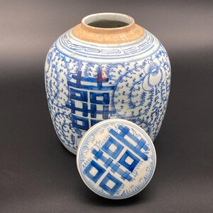 May include: A blue and white ceramic ginger jar with a matching lid, featuring a double happiness symbol and intricate vine patterns. The jar has a wide, rounded body and a tan-colored unglazed rim at the opening, with the lid placed in front.