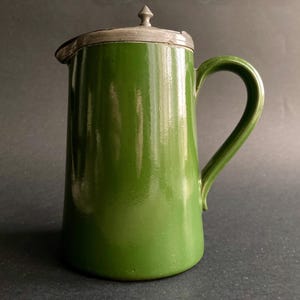 May include: A green enamel pitcher with a silver-coloured lid and handle. The pitcher has a glossy finish and a classic design, suitable for serving beverages. The handle is curved, and the lid has a small decorative finial.