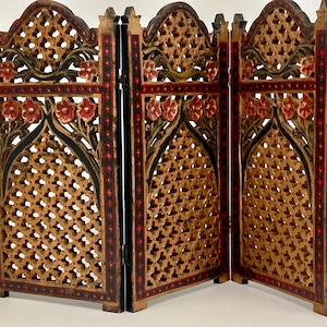 Könnte beinhalten: Ein dreiteiliger Faltbildschirm mit aufwendigen Gitterholzarbeiten und geschnitzten Blumenmotiven. Das Holz ist in einem Naturton gehalten und mit schwarzen und roten Rändern sowie Akzenten versehen. Jedes Paneel hat einen Spitzbogenabschluss.