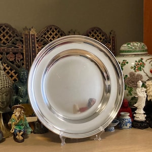 Puede incluir: Un plato circular de metal plateado con una superficie lisa y reflectante y un borde elevado y redondeado. El plato se exhibe en posición vertical sobre un soporte de plástico transparente, mostrando su acabado pulido y su diseño simple y limpio.