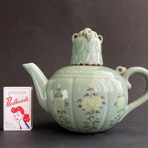 May include: A celadon green ceramic teapot with a matching lid. The teapot features floral designs in yellow and black. A box of Redheads matches is next to the teapot.
