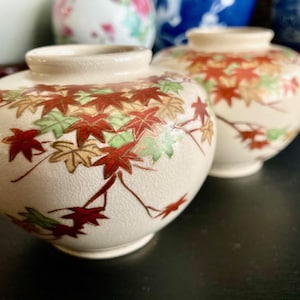 Peut inclure: Deux petits vases en céramique blanche avec un motif de feuilles d'érable rouge, or et vert. Les vases sont ronds et ont une finition brillante.