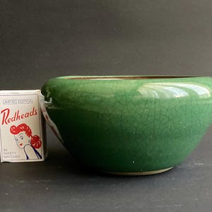 May include: A small, round, green ceramic bowl with a crackle glaze. The bowl is next to a matchbox with the text "Redheads" and an illustration of a woman.