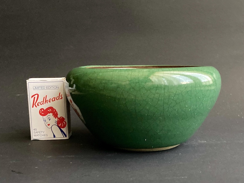 May include: A small, round, green ceramic bowl with a crackle glaze. The bowl is next to a matchbox with the text "Redheads" and an illustration of a woman.