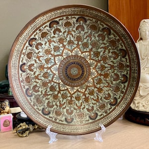 Peut inclure: Grande assiette décorative ronde avec un motif floral détaillé dans les tons marron, or et vert. L'assiette a un bord marron foncé et un motif de médaillon central. L'assiette est présentée sur un support transparent.
