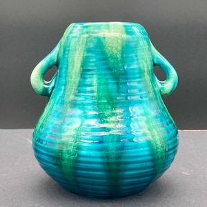 May include: A turquoise and emerald green ceramic vase with two side handles, featuring a ribbed texture and a visible crackle glaze finish. The vessel has a rounded body that tapers towards the top opening, displaying vertical colour drips.