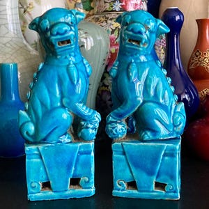 May include: A pair of turquoise ceramic Foo Dog statues. Each dog sits on a square base and holds a ball. The statues have detailed features and a glossy finish. Various vases are in the background.