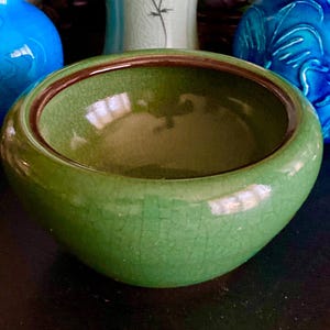 May include: A green ceramic bowl with a crackle glaze and a dark brown rim. The bowl is round and has a wide opening. Two blue decorative vases are in the background.