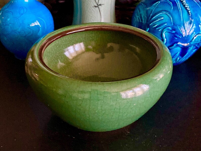 May include: A green ceramic bowl with a crackle glaze and a dark brown rim. The bowl is round and has a wide opening. Two blue decorative vases are in the background.