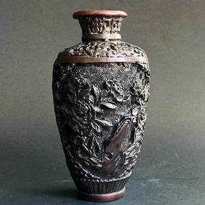 May include: A dark brown, intricately carved vase with a flared base and a narrow neck. The vase features detailed floral and leaf patterns, with a textured surface. The rim and base have a reddish-brown hue, adding contrast to the darker body.