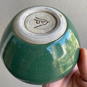 May include: A handcrafted, green ceramic bowl with a crackle glaze. The bowl's base is a lighter color with a stamped design. The bowl is held in a hand.